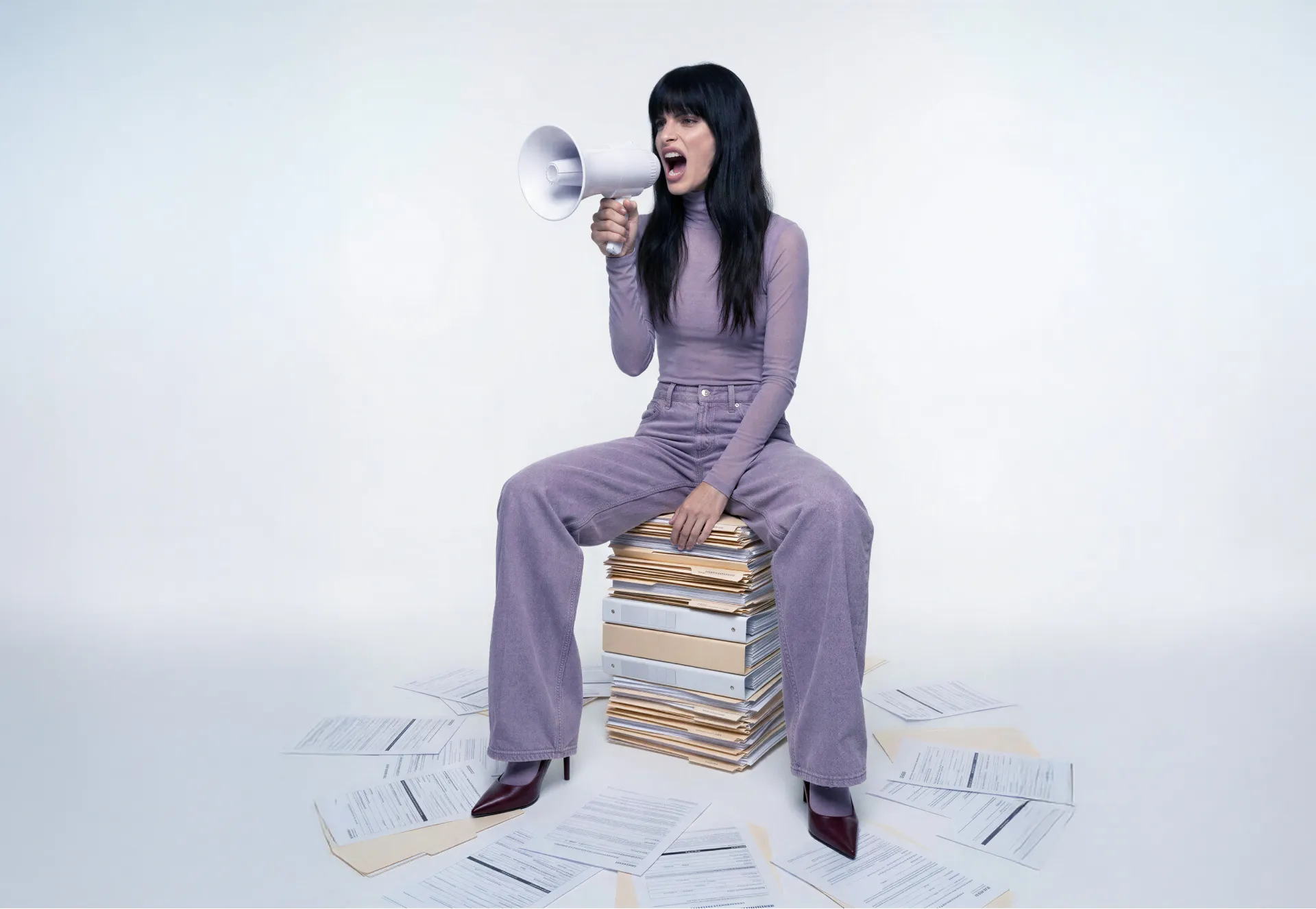 Illustrationsbild zum Artikel Arbeitgeberzuschuss bAV: Frau in lilafarbenem Outfit sitzt auf einem Stapel Aktenordner und ruft mit einem Megafon — rund um sie herum verstreute Dokumente auf dem Boden.