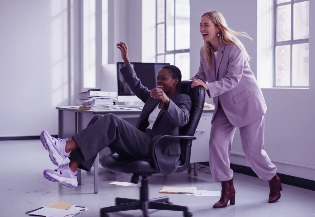 Zwei Frauen lachen im Büro, während eine die andere auf einem Bürostuhl durch den Raum schiebt