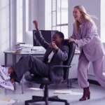 Zwei Frauen lachen im Büro, während eine die andere auf einem Bürostuhl durch den Raum schiebt